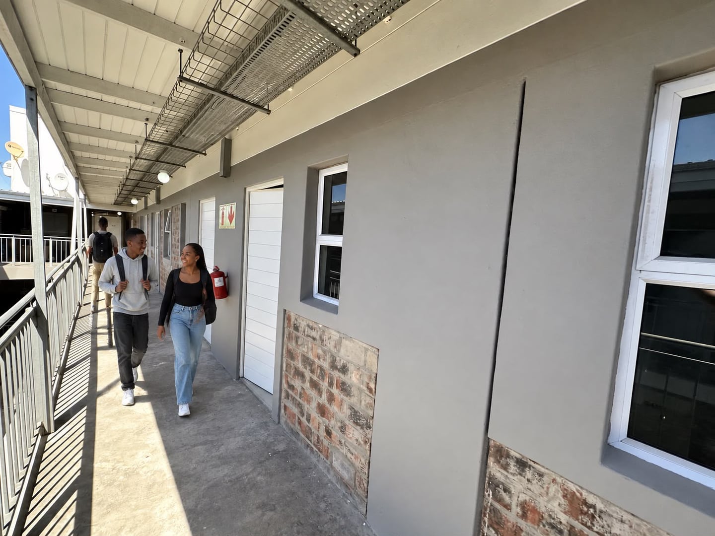 People walking along a covered, open-sided apartment passage with metal balustrades and doors beside exposed-brick wall panels.